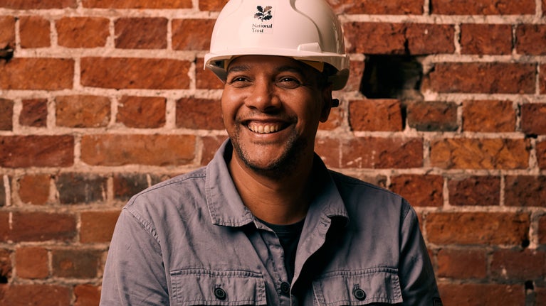 Picture of artist Rich Miller smiling wearing a National Trust hard hat with red brickwork in the background.